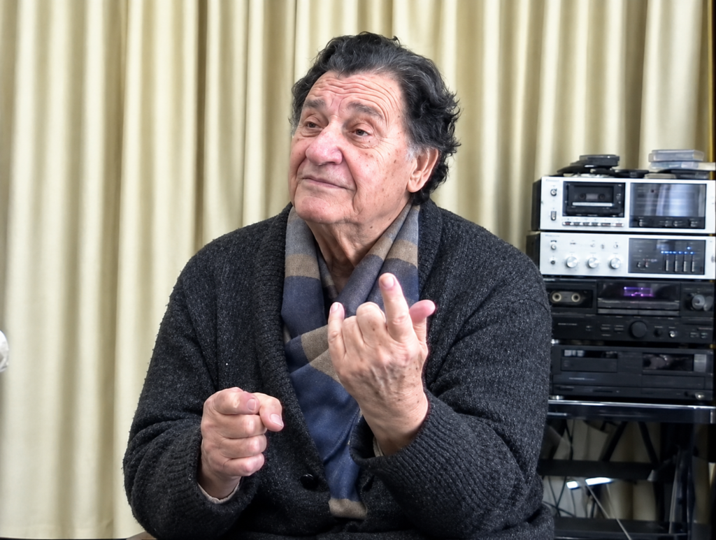 An older man in a dark cardigan and scarf speaking, seated in front of a curtain and vintage audio equipment